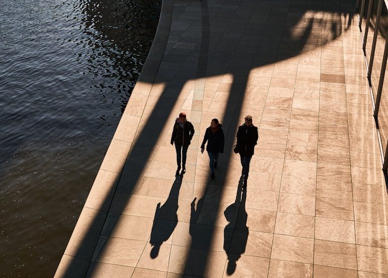 Drei Personen gehen entlang einer Uferpromenade, ihre Schatten sind zu sehen.